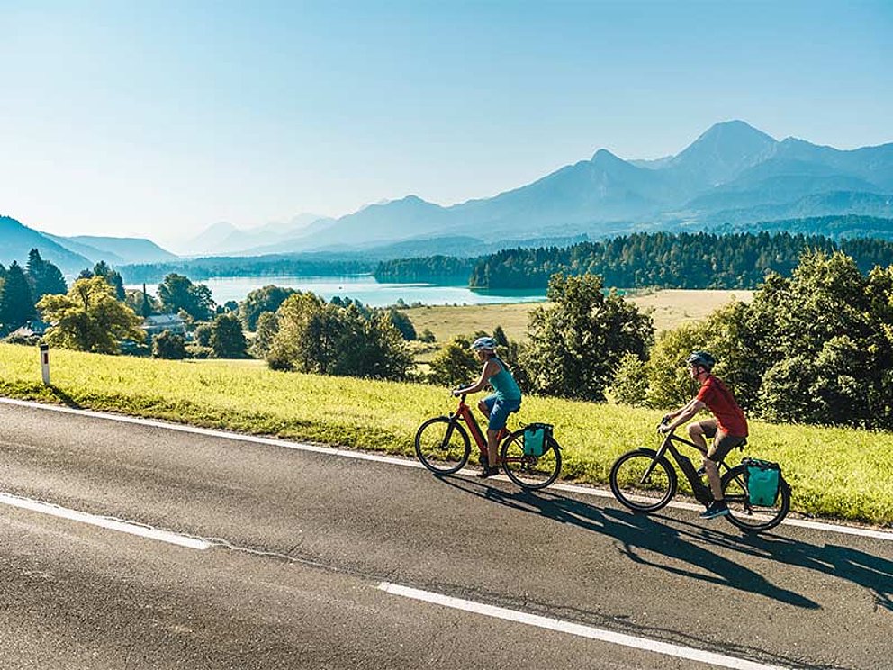 2 Radfahrer auf der STraße. Im Hintergrund ein herrliches Panorama mit Seen und Bergen