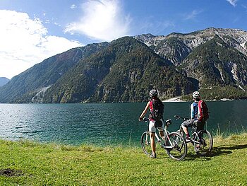 2 Mountainbiker stehen am Ufer eines Bergsees. Im Hintergrund alpine Bergwelt.