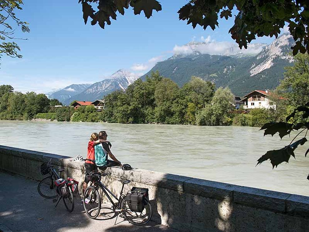 Zwei Radfahrer genießen bei Sonne die tolle Aussicht auf den Fluss und die Berge