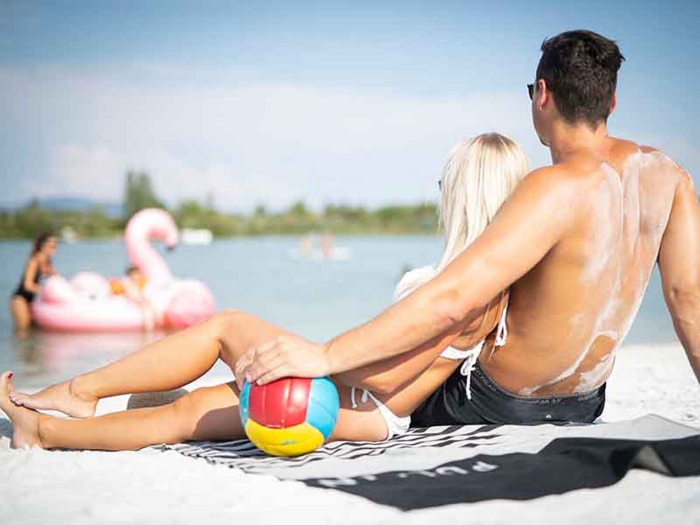 Junges Paar am Seeufer mit Wasserball. Im Wasser ein Mutter mit Kind und Schwimmtier