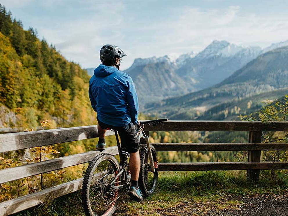 Ein Mountainbiker von hinten an ein Geländer gelehnt, der die Aussicht in die Berge genießt.