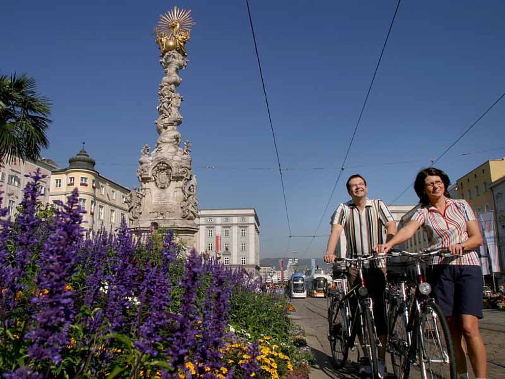 Radfahrer schieben die Fahrräder über den Hauptplatz in Linz, vorbei an der Pestsäule und Blumenbeeten.