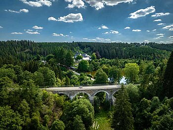 Ein ehemaliges Eisenbahn Viadukt, auf dem Radfahrer radeln. Wiesen und Wälder sowie ein Fluss sind zu sehen.