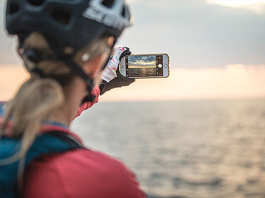 ein Dame mit Radhelm macht ein Foto mit ihrem Smartphone in Richtung Meer