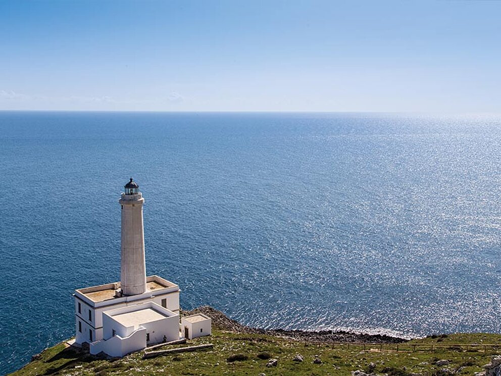 Ein strahlend weißer Leuchtturm auf einer Klippe über dem tiefblauen, glitzernden Mittelmeer in Apulien, Italien.
