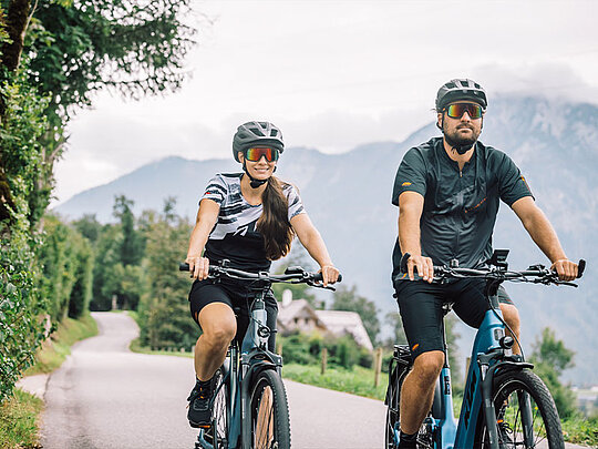 ein Paar auf KTM-E-Bikes auf einem asphaltierten Radweg, im Hintergrund Berge.