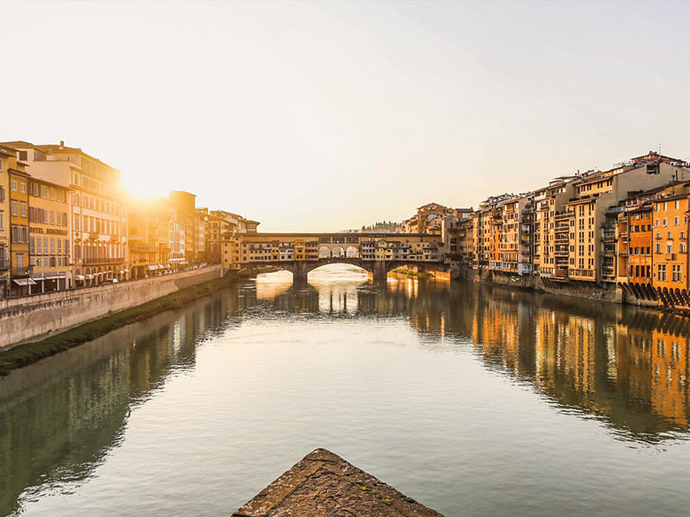 Blick auf die berühmte Brücke Ponte Vecchio in Florenz bei Sonnenuntergang, gespiegelt im Fluss Arno.