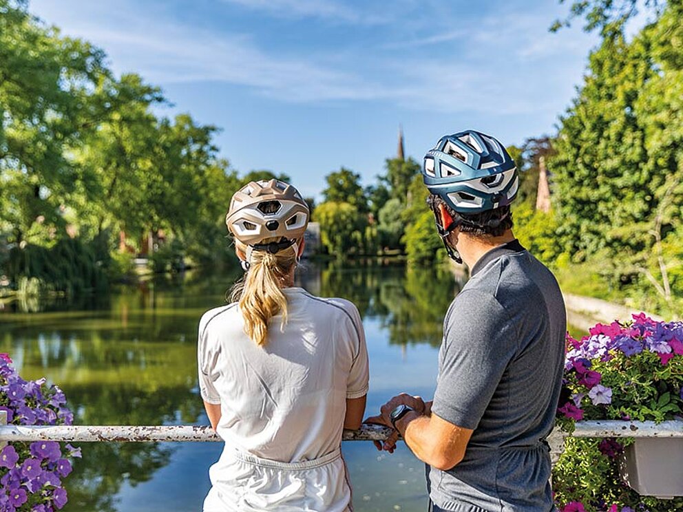 Zwei Radfahrer mit Helmen blicken von einer Brücke aus auf einen ruhigen Kanal in der belgischen Natur.