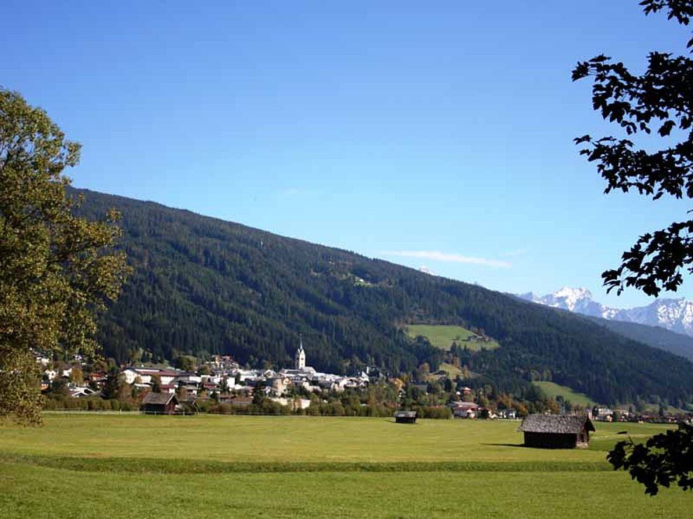 Blick auf Radstadt nahe dem Ennsradweg. Der Ort liegt in einer Ebene mit grünen Wiesen, dahinter ein bewaldeter Bergrücken.