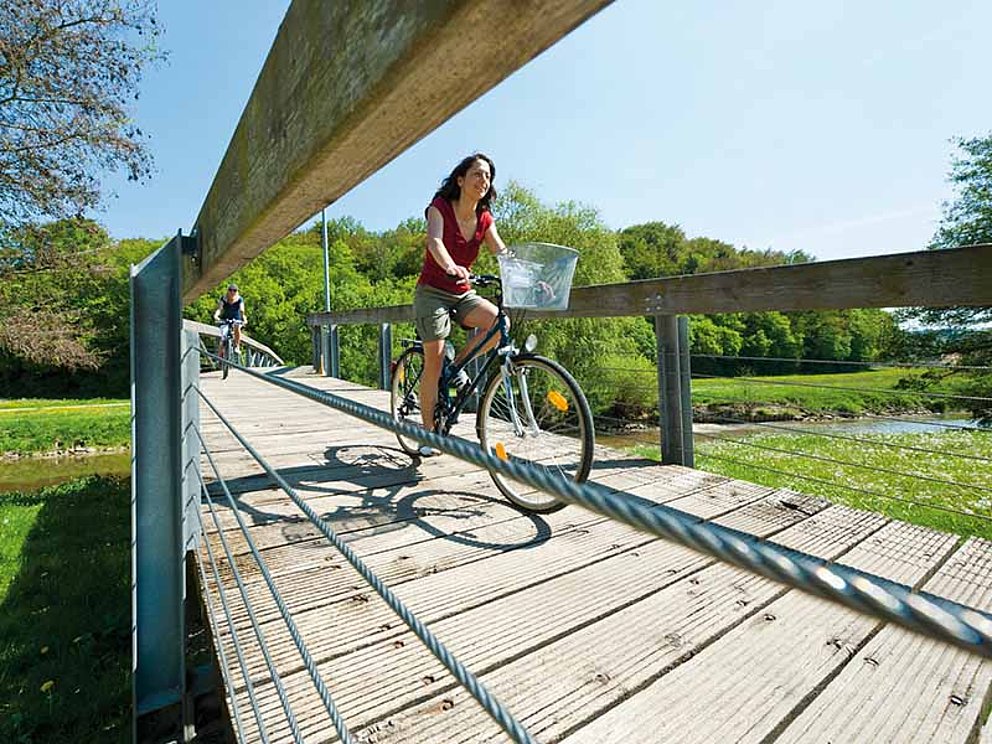 female cyclist crosses a bridge, river and meadows in the back