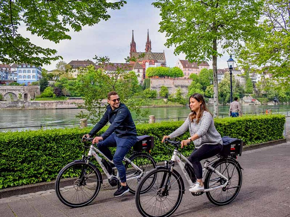 2 Radfahrer am Ufer des Rhein, im Hintergrund Stadtpanorama von Basel