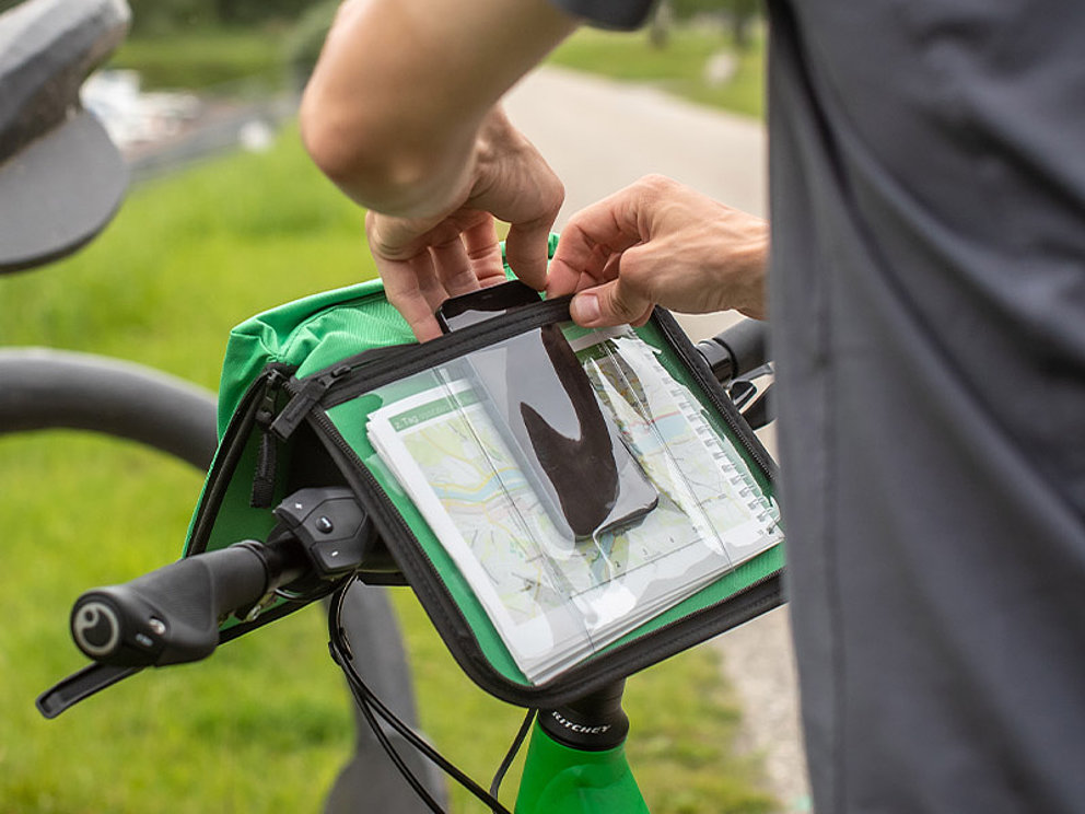 Eine Person steckt das Smartphone mit der Navi-Anwendung in die dafür vorgesehene Tasche in der Lenkertasche am Fahrrad