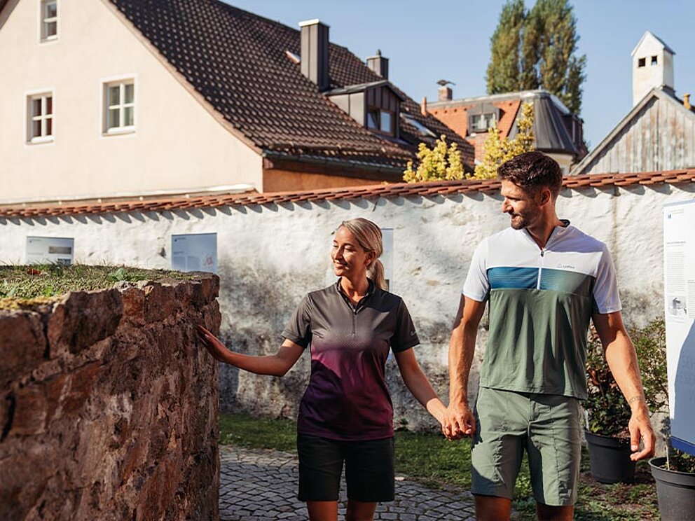 Ein Paar hält Händchen an einer historischen Steinmauer des Kastell Biotro im Römermuseum in Passau während einer Radreise auf dem Donauradweg.