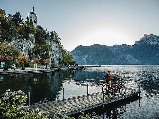 2 Personen mit Bikes am Steg am See, im Hintergrund Traunkirchen