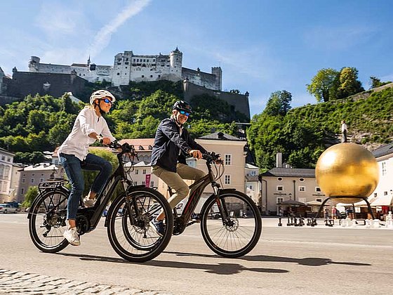 Mann und Frau auf E-Bikes am Mozartplatz in Salzburg, dahinter die Festung