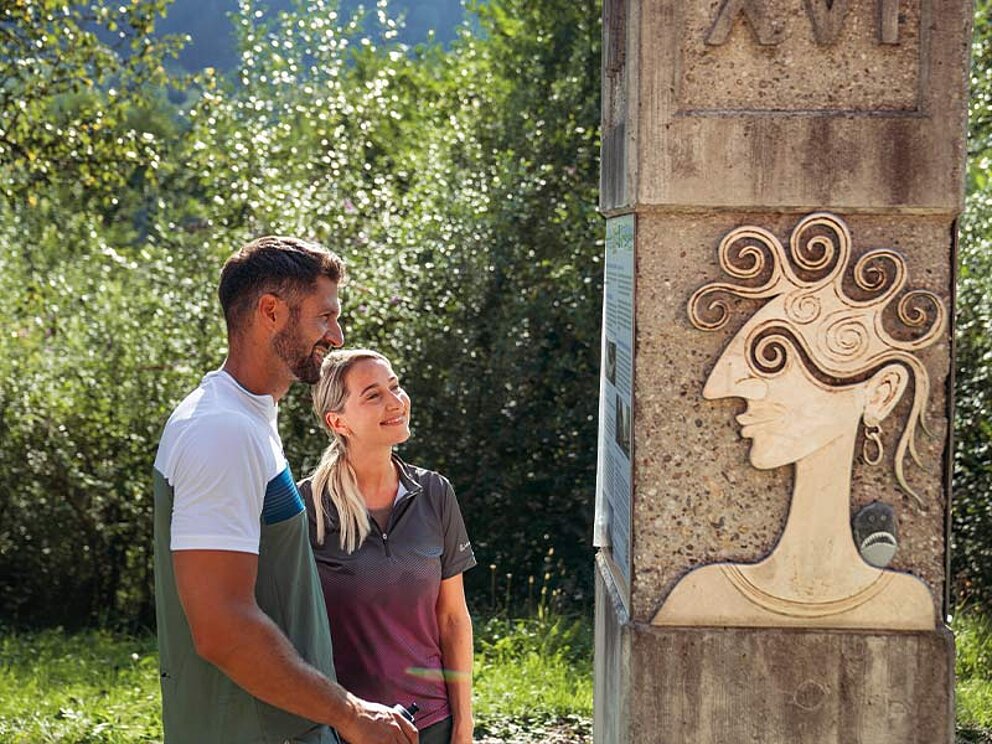 in Paar betrachtet eine römische Stele mit einem geschnitzten Gesicht am Donaulimes in Freizell während einer Radreise auf dem Donauradweg.