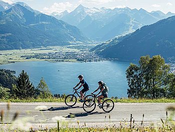 2 cyclists on a road high above Lake Zeller See, mountains in the back