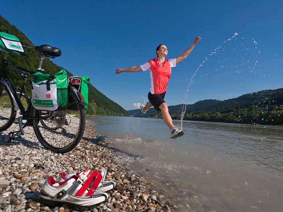 Eine junge Frau springt mit gestreckten Beinen über den Fluss. Ein kleiner Bogen Spritzwasser saust in die Höhe. Am Flussufer steht ein Fahrrad.