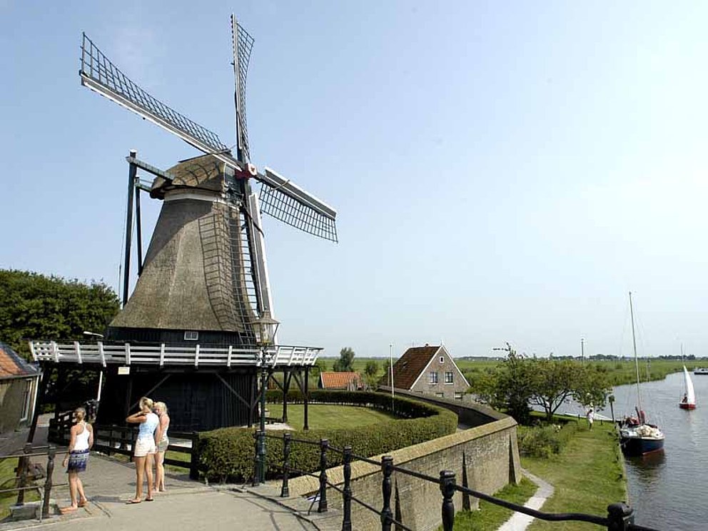 Eine reetgedeckte Windmühle mit 4 Flügeln am Ufer eines Flusses in Holland.