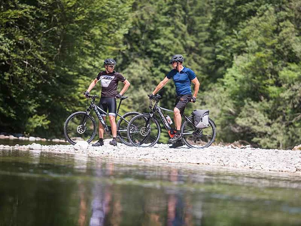 2 cyclists on a riverbank, wood in the back