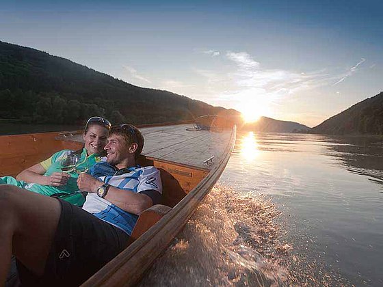 Ein junges Paar liegt im Bug einer Holzzille mitten auf der Donau. Dahinter reflektiert sich die untergehende Sonne im Wasser.