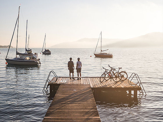 2 cyclists on a stage. boats on the lake, mountains in the far.