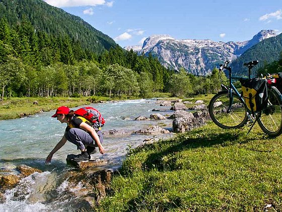 Ein Radfahrer hält eine Rast an der Enns. Er pritschelt im seichten Wasser des Flusses. Das Fahrrad steht in der Wiese am Ufer. Am anderen Ufer erheben sich hohe bewaldete Berge.