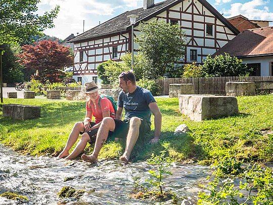 2 Personen sitzen am Ufer eines Bachs und halten die Füße ins Wasser