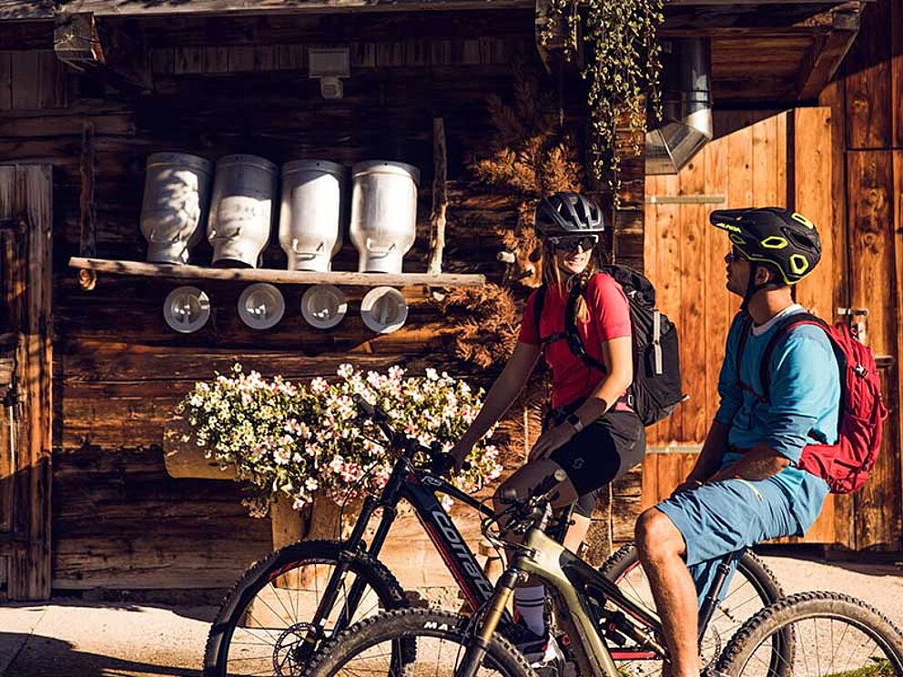2 Mountainbiker vor einem Holzhaus. Milchkannen trocknen in der Sonne