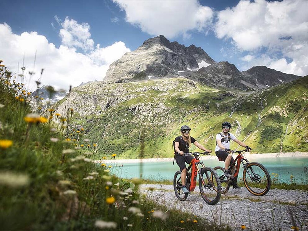 2 Mountainbiker vor einem Bergsee, im Hintergrund Berge