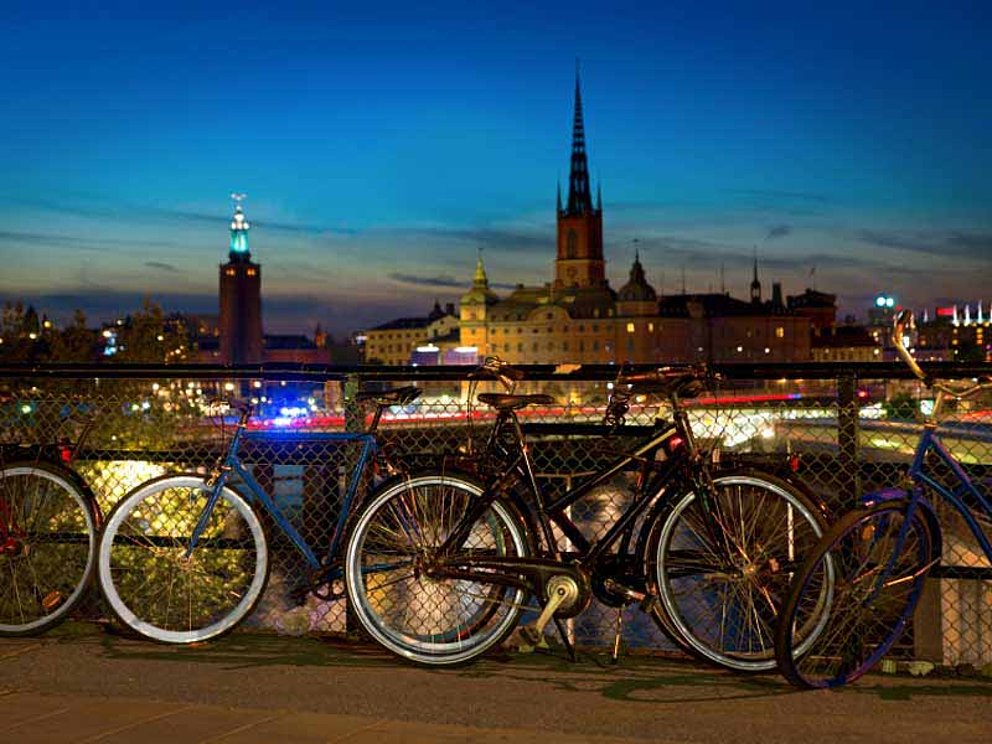 Fahrräder lehnen an einem Geländer vor der beleuchteten Abendkulisse in Stockholm.