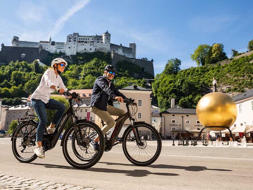 Paar auf E-Bikes am Mozartplatz in Salzburg, dahinter die Festung Hohensalzburg