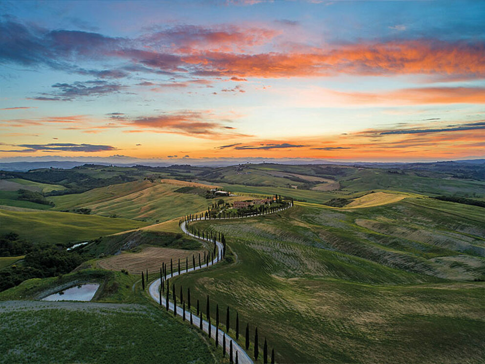  Panorama des Val d'Orcia in der Toskana bei Sonnenuntergang mit einer charakteristischen Zypressenallee, die zu einem Landhaus führt.