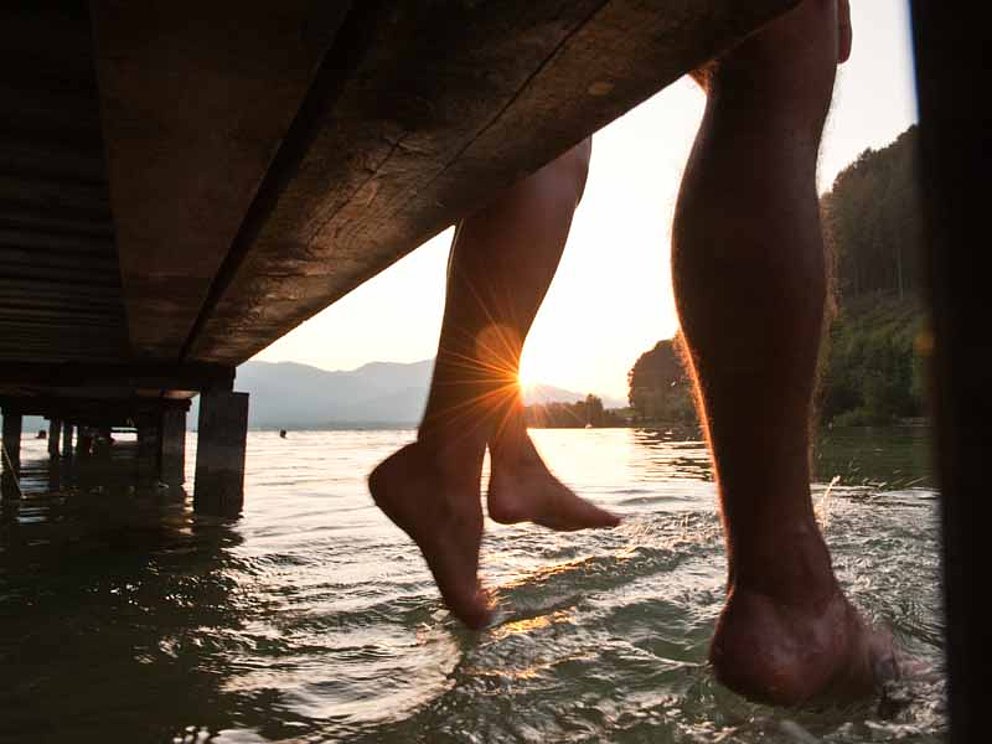Die nackten Füße zweier Personen baumeln von einem Steg. Darunter plätschert der See. 