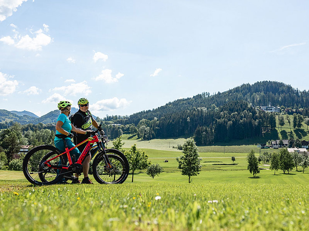Ein älteres Paar mit E-Bikes macht Rast inmitten grüner Wiesen und Hügel