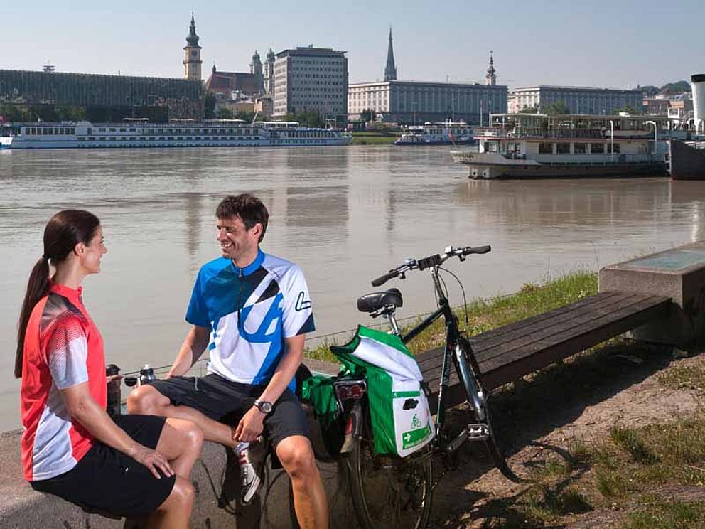 Ein junges Paar in Raddressen sitzt auf einer niedrigen Mauer am Donauufer in Linz. Rechts von ihnen steht ein Fahrrad, im Wasser liegt ein Schiff. Am anderen Ufer winkt die Silhouette der Stadt.