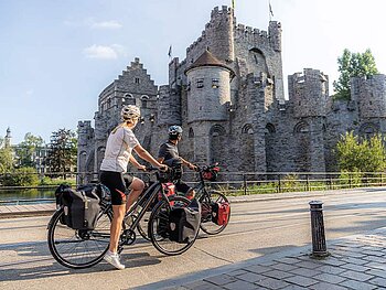 Zwei Radfahrer mit vollgepackten Trekkingrädern und Radtaschen vor der mittelalterlichen Burg Gravensteen in Gent, Belgien.