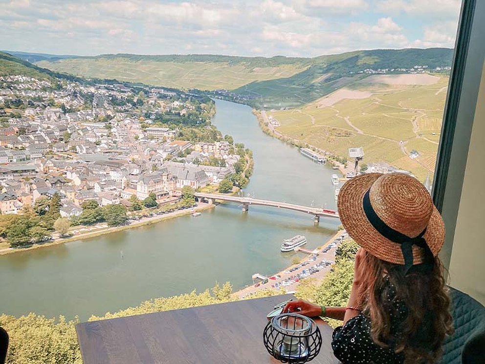 Eine Dame mit Hut sitzt am Panoramafenster auf der Burg Landshut und blickt über die Stadt Bernkastel-Kues und die Mosel.