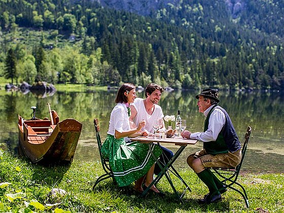 3 Personen in Tracht sitzen am bewaldeten Seeufer und trinken Wein