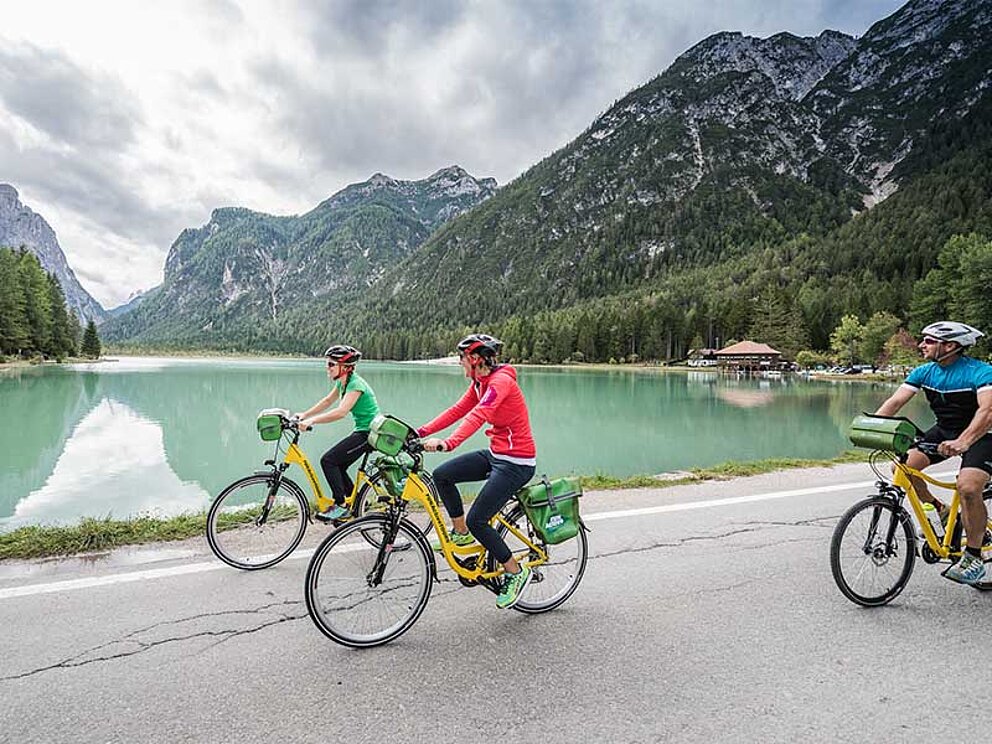 3 Radfahrer auf einem asphaltieren Weg am Ufer eines Sees