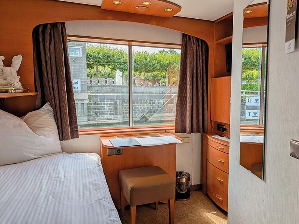 Kabine mit Bett, kleinem Schreibtisch und Schranknische. Ein Fenster ist ebenfalls vorhanden.
