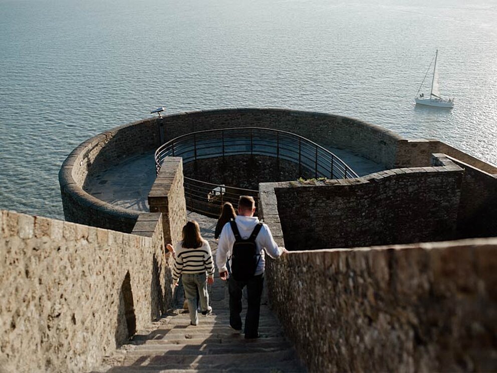 2 Personen gegen stufenabwärts auf der Festung, im Hintergrund Meer und ein Segelboot