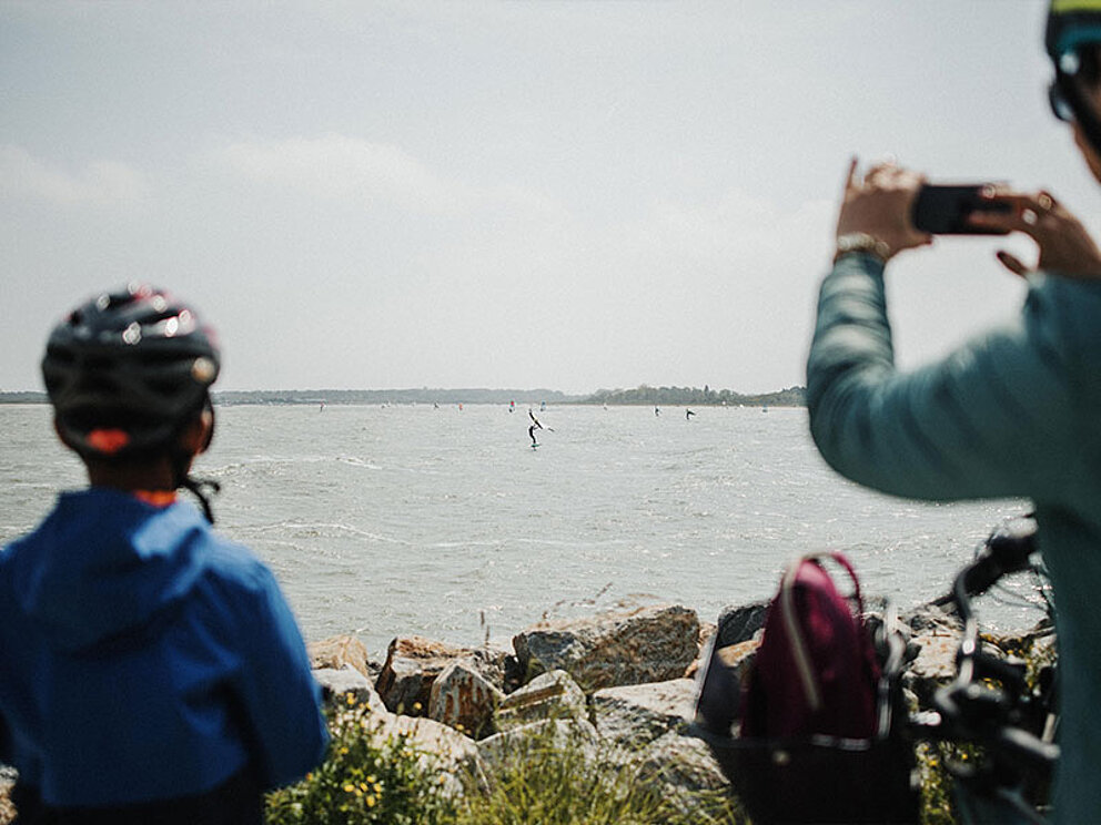 2 Personen mit Radhelm und Handy von Hinten, im Hintergrund das Meer mit Windsurfern