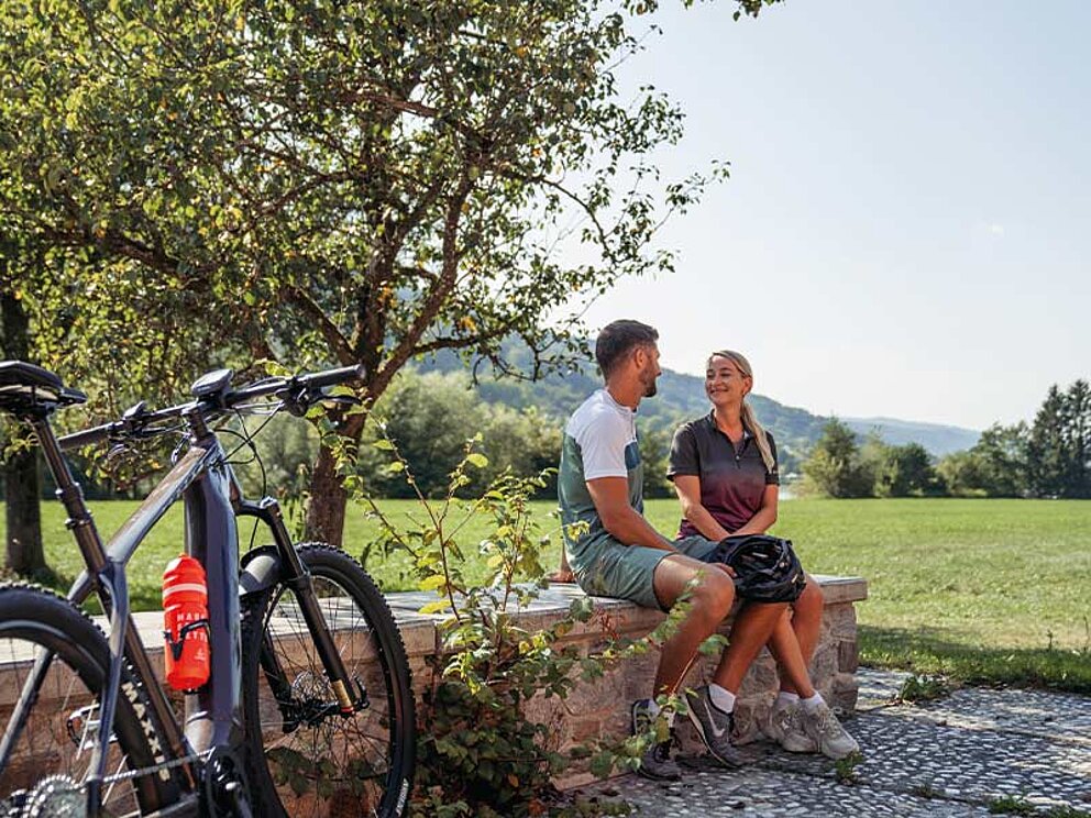 Ein Paar macht eine Rast auf einer Steinmauer bei ihrer Radtour am Donaulimes in Freizell; im Vordergrund steht ein modernes E-Bike.