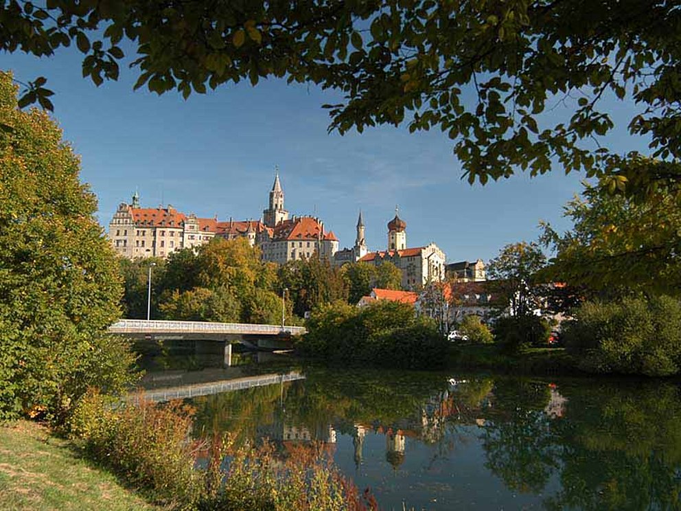 Blick auf Schloss Sigmaringen am Donauradweg. Das Schloss thront auf einer Anhöhe über der Stadt. Im Vordergrund spannt sich eine Brücke über den Fluss. Beidseits an den Ufern wachsen viele Bäume und Büsche.