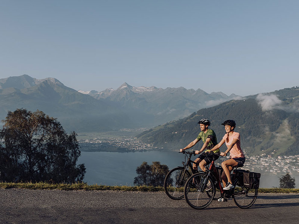 2 Radfahrer am asphaltierten Radweg, im Hintergrund der Zeller See und Berge