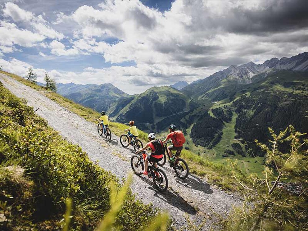 4 Biker fahren bergauf am Schotterweg, umgeben von Bergen, bewölkter Himmel