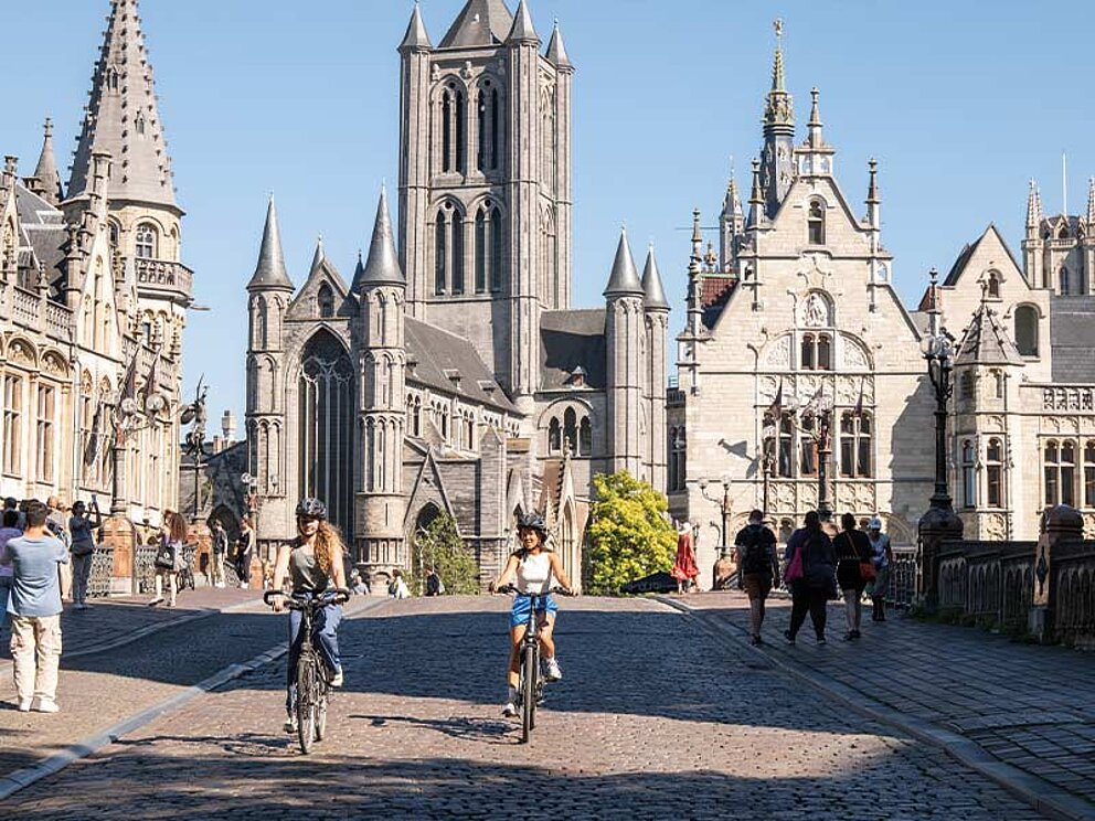 Zwei Frauen radeln über ein Kopfsteinpflaster im historischen Zentrum von Gent, Belgien, vor einer beeindruckenden gotischen Kirche.