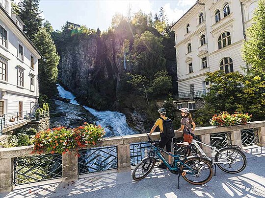 2 Personen mit Fahrrad stehen am Geländer beim Wasserfall im Zentrum von Bad Gastein