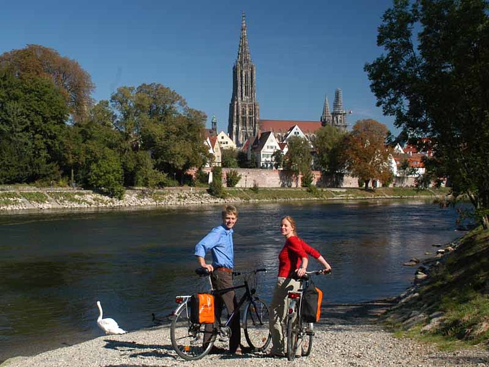 Ein Mann und eine Frau halten mit ihren Fahrrädern am Donauufer. Neben ihnen sitzt ein Schwan. Am anderen Ufer grüßt das Ulmer Münster.